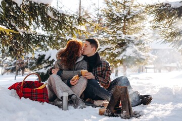 A married couple had a picnic in a snowy forest. Young people sit in front of a small fire and eat pancakes.
