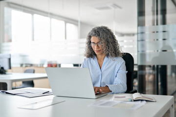 Serious busy mature middle aged professional business woman, older lady manager executive leader wearing glasses looking at laptop using computer in office working on digital project sitting at desk.