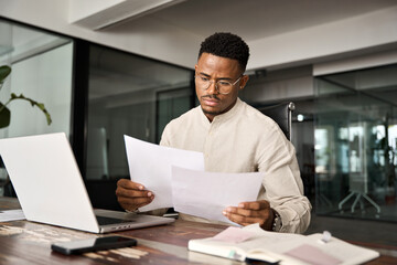 Serious businessman accountant expert reading legal paper company file overview at workplace. Young busy African professional business man checking document working at laptop computer in office.