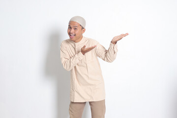 Portrait of shocked Asian muslim man in koko shirt with skullcap showing product and pointing with his hand and finger to the side. Advertising concept. Isolated image on white background