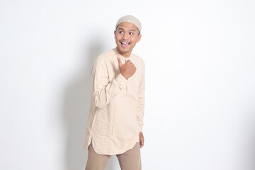 Portrait of shocked Asian muslim man in koko shirt with skullcap showing product and pointing with thumb up to the side. Advertising concept. Isolated image on white background