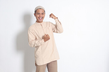 Portrait of religious Asian muslim man in koko shirt with skullcap raising his fist, celebrating...