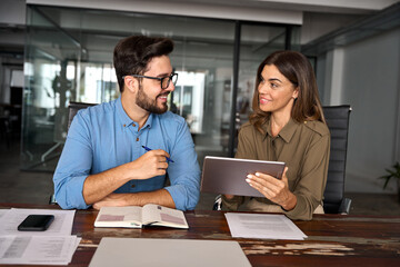 Two busy happy professional business people working in office with digital tablet. Middle aged female executive manager talking to male colleague having conversation sitting at workplace. - obrazy, fototapety, plakaty