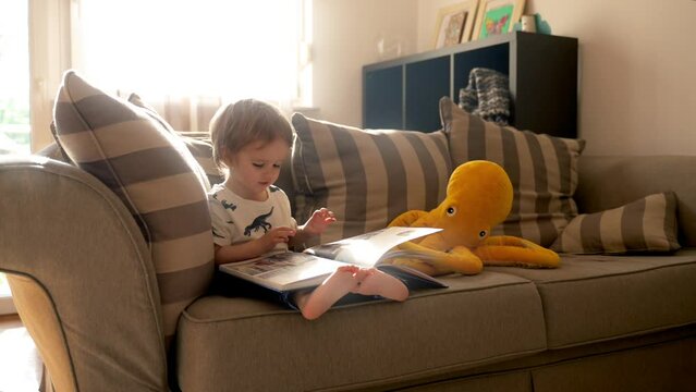 A little boy sits on a couch, engrossed in reading a book.