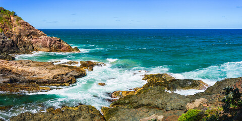 Australian coast, view from a cliff to the blue ocean with a rocky shore on a sunny day. Sea landscape, waves crashing on volcanic rocks at the shore.