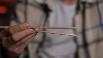 Close-up cropped shot of unrecognizable man practicing chopsticks in Japanese restaurant. Closeup hands of Caucasian male trying to eat sushi rolls or noodles using chopsticks. Shooting in slow motion