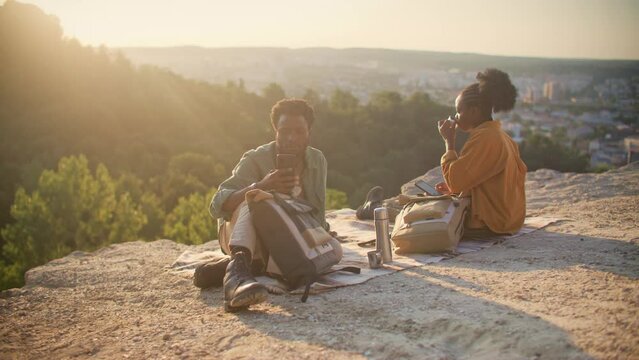 Close view of African American couple sitting on blanket at edge of cliff and enjoying their meal. While resting checking mobile devices to check with family. Using smartphones to stay in contact.
