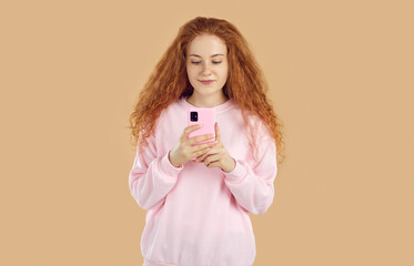 Calm, interested, relaxed redhead pretty curly girl in pink sweatshirt is looking at smartphone screen, reading message or watching video, talking video chat on beige background. Online life concept.