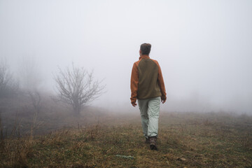 Naklejka na ściany i meble Back view of young girl hiking traveling going on top of cliff in travel, mountaineer girl goes into the fog, tourist in a foggy field far from civilization, foggy future, without a path