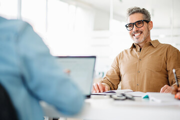 Mature grey-haired professional man in a tan shirt smiles engagingly at a colleague during an...