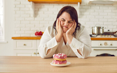 Young fat upset overweight woman sitting at the table in kitchen looking at the plate with sweet donuts grimacing with unhappy face expression at home. Diet restrictions, losing weight concept. - obrazy, fototapety, plakaty