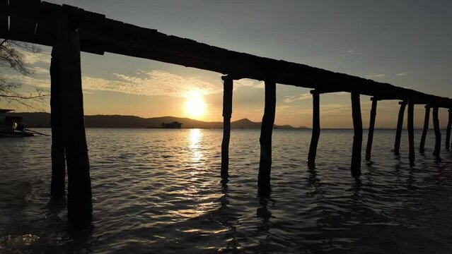 Serene sunset view under a wooden pier with calm sea waters and a silhouette of hills in the background capturing a tranquil evening moment