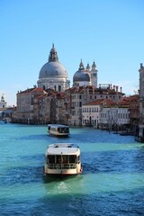 Panorama sur la ville de Venise, avec un bateau (vaporetto) naviguant sur l’eau bleue du Grand Canal et vue sur le dôme de la basilique Santa Maria della Salute dans le quartier du Dorsoduro (Italie)
