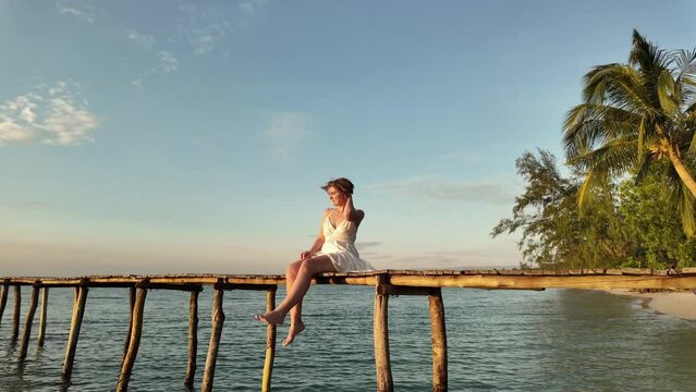 Woman sitting on a wooden pier by the ocean at sunset with tropical palm trees in the background evoking a serene vacation vibe