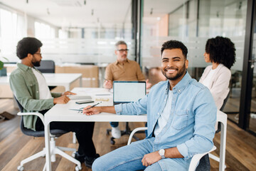 The focus is on a smiling young Arab professional in a denim shirt, sitting in front of a modern...