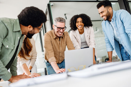 A cheerful group of diverse coworkers lean in around a laptop, indicative of an interactive tech discussion and the high spirits characteristic of a productive and forward-thinking team.