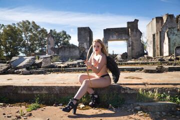 Fototapeta premium Young, beautiful, blonde woman with black angel wings, with the paint on her eyes smeared, with bow and arrow, sitting in the middle of some ruined buildings. Concept fallen angel, lack of love.
