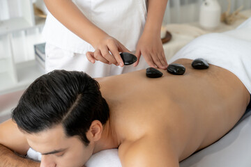 Obraz na płótnie Canvas Hot stone massage at spa salon in luxury resort with day light serenity ambient, blissful man customer enjoying spa basalt stone massage glide over body with soothing warmth. Quiescent