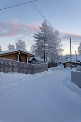 Fototapeta premium Snow-covered street with fences in the village. Log houses, trees, poles, power lines. All in the snow. Day during the polar night in the Russian Far North.