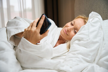 Smiling lady using cellular phone in bedroom upon morning awakening