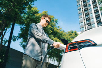 Young man travel with EV electric car charging in green sustainable city outdoor garden in summer...