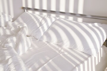 Crumpled white linen on the bed after waking up. Shadows from the blinds on the window fall on the bed.