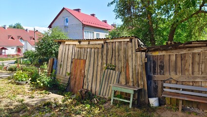 Under the trees stands an old building - a shed made of boards and covered with slate. There is grass growing near it and a table and bench. Further on there are brick buildings with metal tile roofs