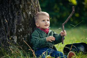 Little Cute Toddler with Wooden Stick Sitting by the Tree - 726714488