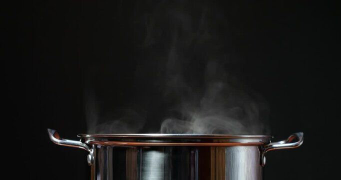Steaming hot water in a steel pan. Cook lunch and dinner in the kitchen. Steam rises from boiling water against a black background. Cooking dinner from porridge or pasta broth.