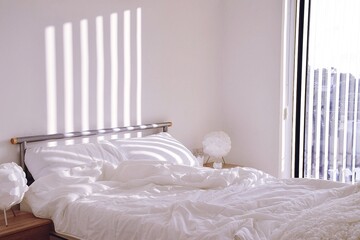 Crumpled white linen on the bed after waking up. Shadows from the blinds on the window fall on the bed.