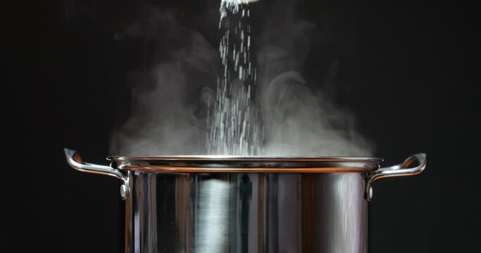 Steaming hot water in a steel pan. Salt the water with your hand or a spoon. Cook dinner and food in the kitchen by adding seasoning. Steam rises from boiling water against a black background.