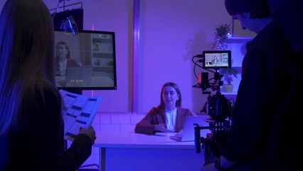 The crew backstage. Female producer, videographer and female presenter in a dark studio in blue and pink neon lights during filming. A female producer with storyboard sheets in her hands.