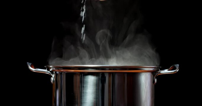Steaming hot water in a steel pan. Salt the water with your hand or a spoon. Cook dinner and food in the kitchen by adding seasoning. Steam rises from boiling water against a black background.