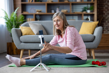 Senior mature gray haired woman doing fitness sitting on exercise mat at home in living room on...