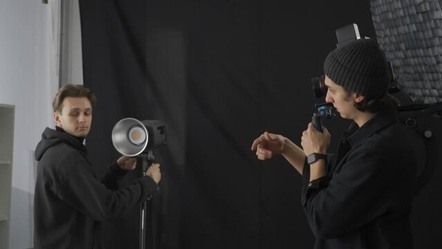 A male cameraman discusses the shooting process with a male lighting technician. The male lighting technician adjusts the lighting in the studio. Backstage video production recording.
