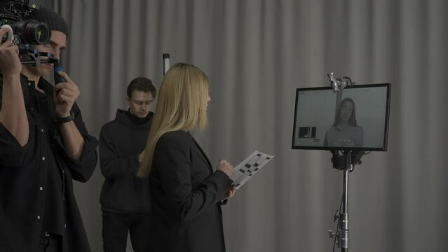 Female director, cameraman and male illuminator in the studio during filming. Female director with a script stands in front of the monitor, nods her head, and gives instructions to the cameraman.