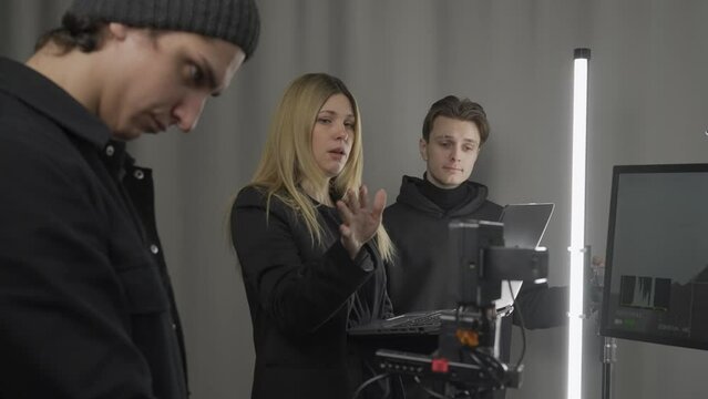 Female producer, camera operator, lighting technician in the studio during filming. The cameraman adjusts the camera, the producer shows the lighting engineer the shooting, lighting plan on her laptop