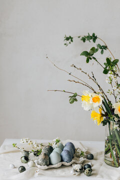 Happy Easter! Stylish easter eggs and spring flowers on linen rustic table. Natural painted marble blue eggs in tray and daffodils bouquet. Modern minimal still life
