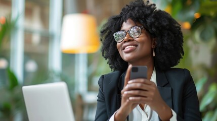 African black business woman using smartphone while working on laptop at office. Smiling mature african american businesswoman looking up while working on phone. Successful woman entrepreneur.