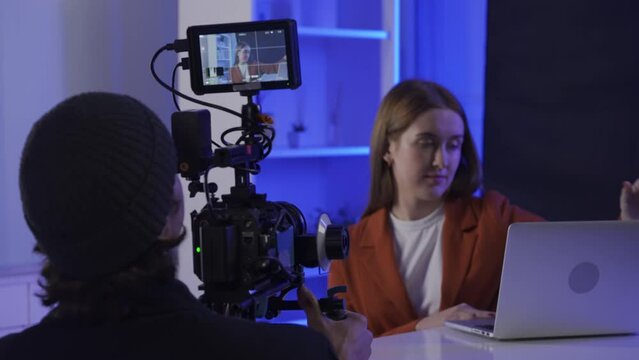 Backstage video production recording. A videographer uses a professional camera to film a female presenter sitting at a table in front of a laptop. Film crew in the studio in blue neon lighting.