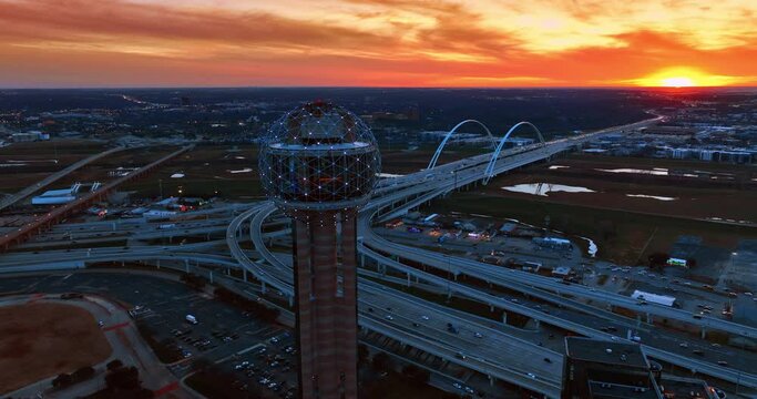 Flying along the spherical top of Reunion Tower in Dallas, Texas, USA at sunset. Complicated road system, Dallas Margaret Mcdermott Bridge and city view at the backdrop of orange sky.