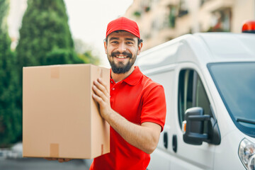Delivery courier service. Delivery man in red cap and uniform holding a cardboard box near a van truck delivering to customer home. Smiling man postal delivery man delivering a package