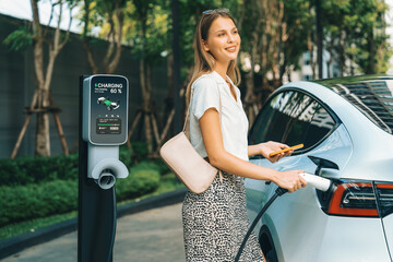 Young woman travel with EV electric car charging in green sustainable city outdoor garden in...