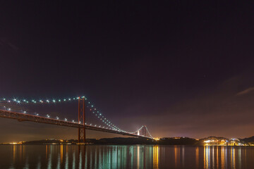 Illuminated red bridge 25 de Abril Bridge crossing the Tagus river with statue of Cristo Rei during night time, Lisbon, Portugal