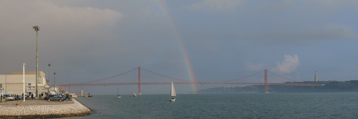 Panorama of rainbow over red bridge 25 de Abril Bridge with sailing boat during sunset, Lisbon, Portugal