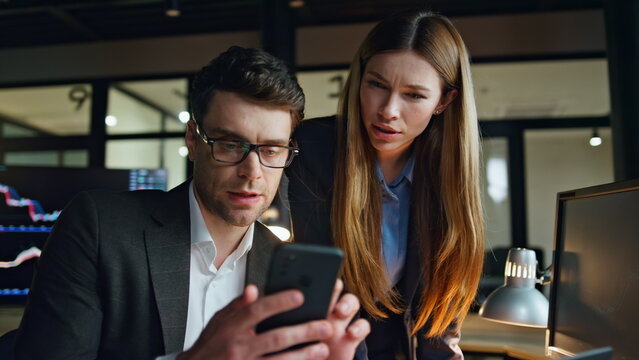 Two business people working dark finance office. Focused man using smartphone