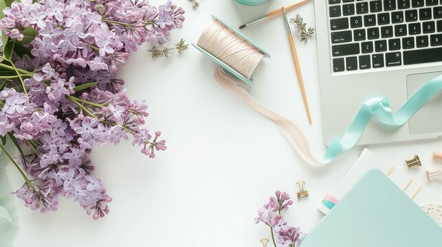 Flat lay, top view office table desk. Workspace with paintbrush, laptop, lilac flowers bouquet, spool with beige and blue ribbon, mint diary on white background.