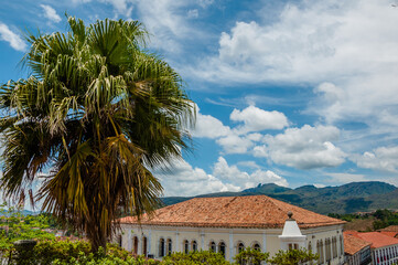 Fototapeta na wymiar The peak of Itacolomi in the background on a day with clouds and sun and in the foreground a palm tree and an old house in the city of Ouro Preto