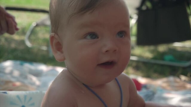 a baby sitting in a kiddie pool on a hot summer day. The baby looks around curiously while the mother pours water on him, making him giggle with delight. It's a precious moment of mother-child bonding
