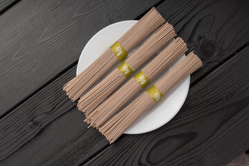 Bundles of brown noodles tied with green paper bands on a white plate over a dark wooden table.